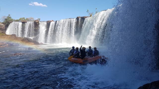 A group of helmeted rafters in an orange inflatable raft navigating churning whitewater directly beneath a wide curtain waterfall on a brazil 4x4 tours expedition, with a rainbow visible in the mist to the left under a clear blue sky