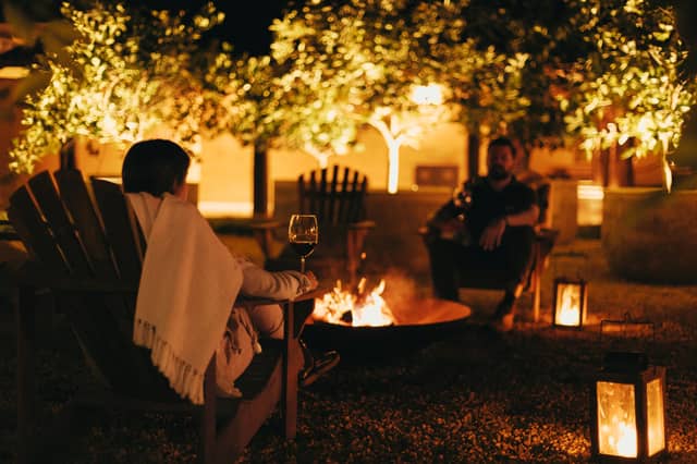 Two guests enjoying brazil luxury tours unwinding by a glowing fire pit at night, wrapped in blankets with red wine and lantern light flickering beneath illuminated trees