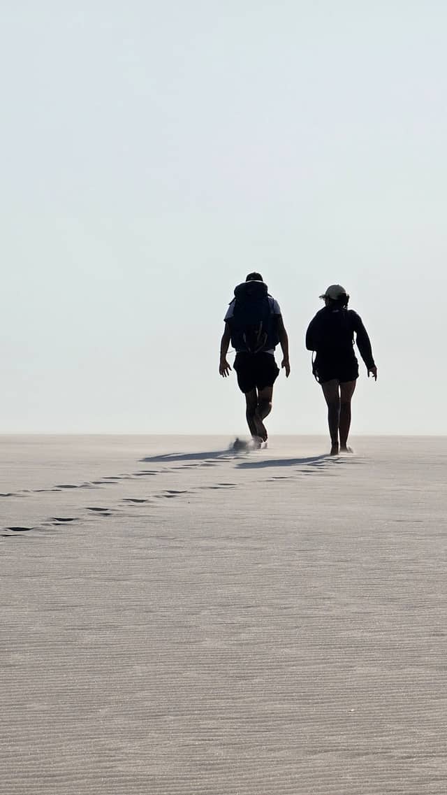 Two trekkers with backpacks walk across a vast, wind-rippled sand flat, leaving a trail of footprints behind them during a multi-day trek in Brazil