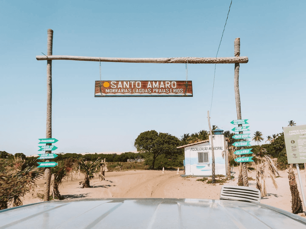 Rustic portico with a wooden sign displaying the text Santo Amaro Morvanas Lagoas Prajasthios over a sandy road.