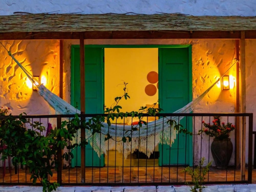 Intimate balcony lit at night with a resting hammock stretched in front of the green double door of a Quintal das Areias chalet.