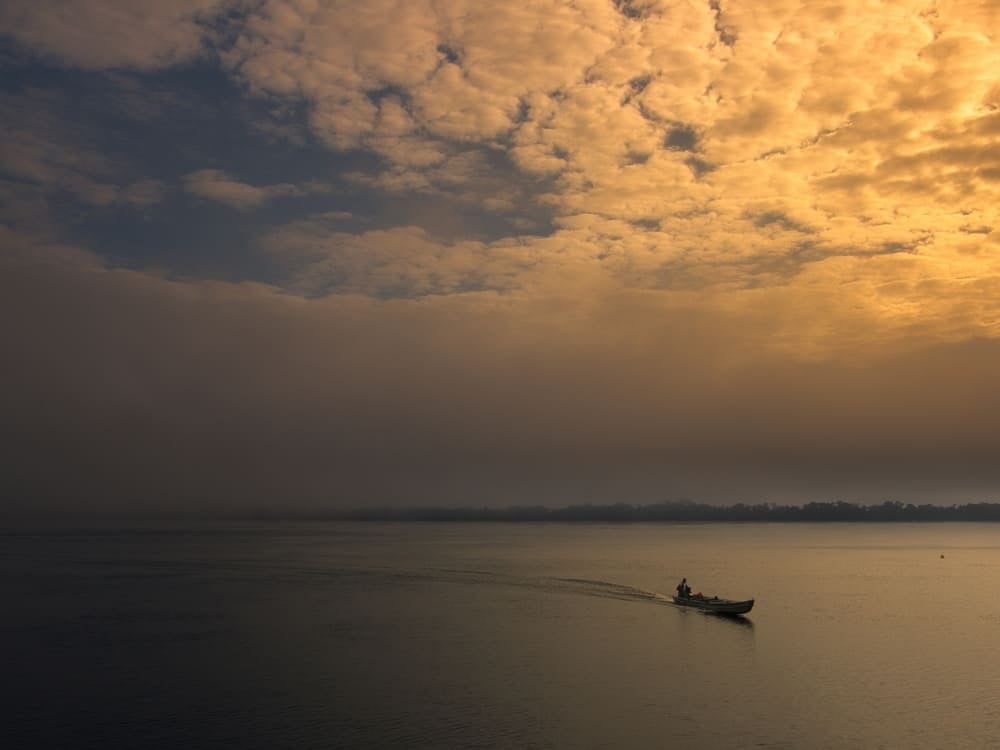 A small motorized canoe traveling across the wide Amazon River during a golden sunrise under a cloudy sky.