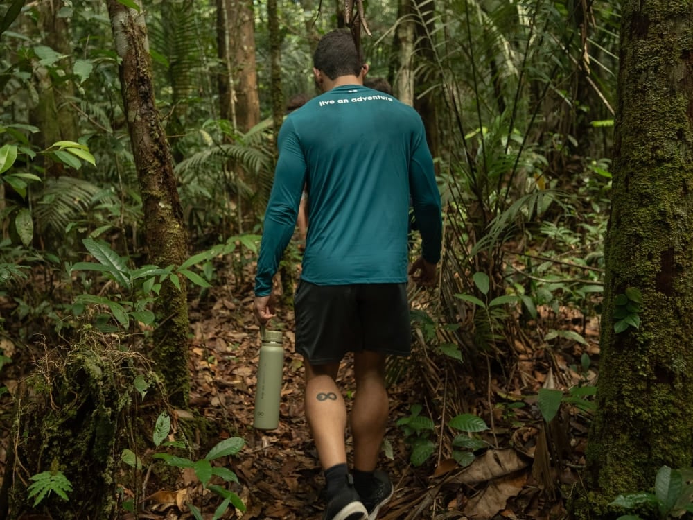 A traveler carrying a reusable water bottle hikes through a lush green Amazon Rainforest trail.