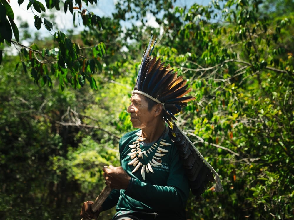 An Indigenous man wearing a traditional feather headdress walks through the lush green Amazon jungle.
