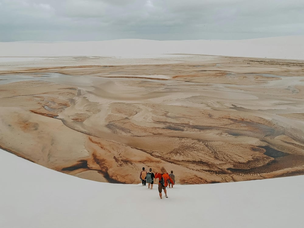 A group of tourists walks across a dry, brown lagoon bed surrounded by large white sand dunes during the dry season.