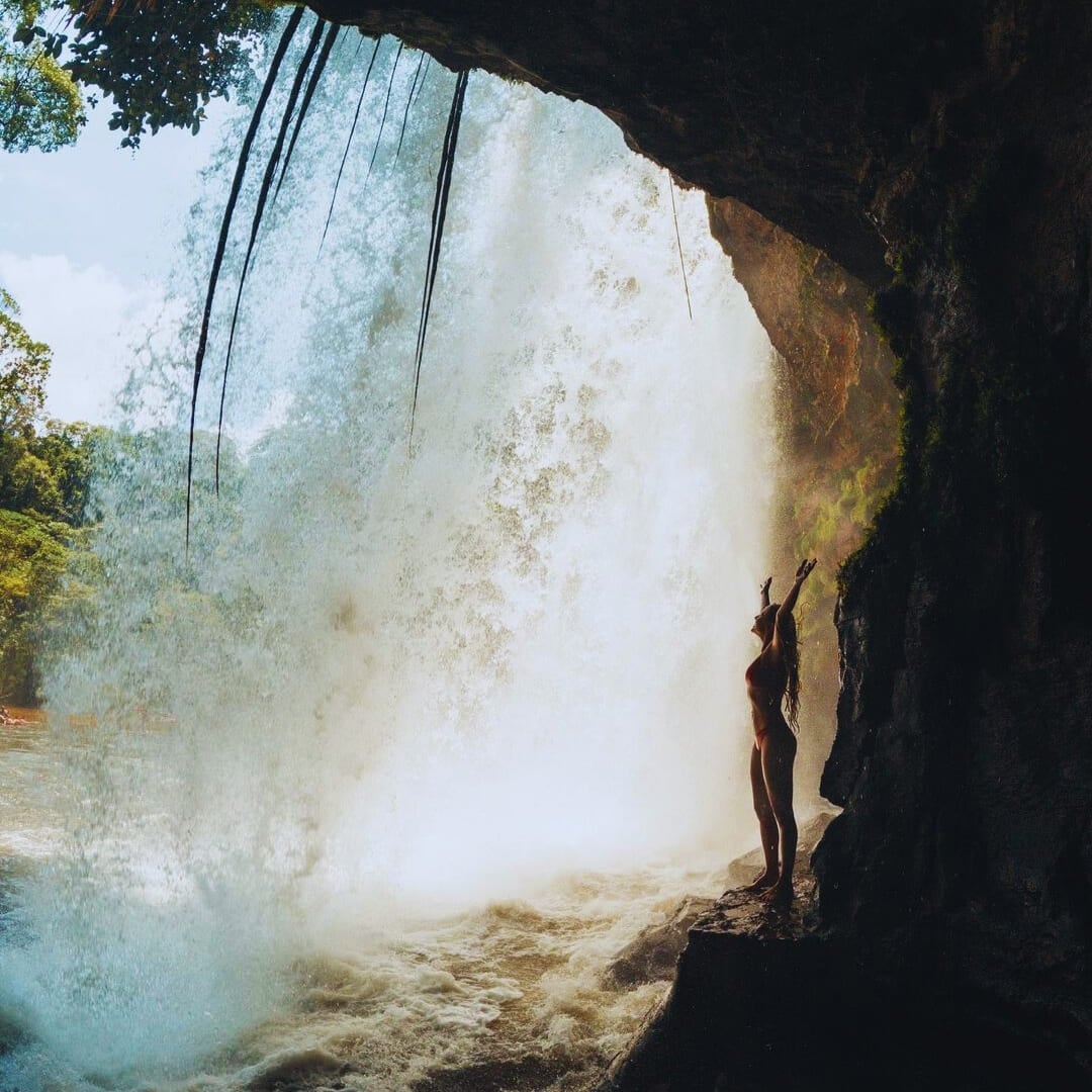 A traveler stands behind a powerful rushing waterfall in the Amazon Rainforest with their arms raised.