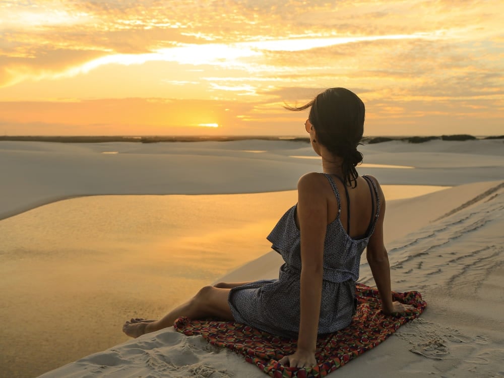 Woman sitting on a beach sarong on the white sand, admiring the golden sunset reflecting on the waters of the Lençóis Maranhenses lagoons.