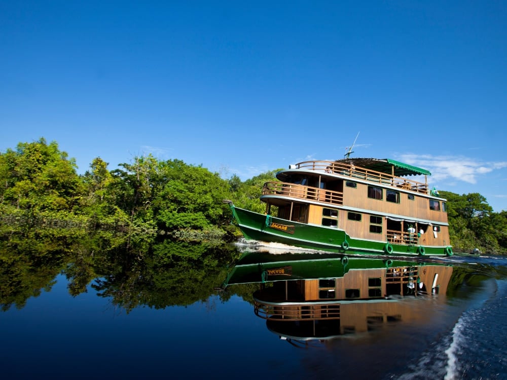 A rustic, three-deck wooden riverboat cruises smoothly along a dark Amazonian river beneath a blue sky.