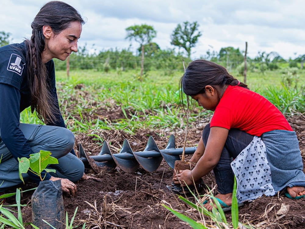 A woman from the SOS Pantanal project and a young local girl use an earth auger to plant a tree seedling together.