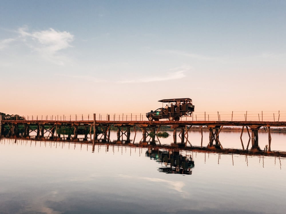 A safari vehicle crosses a wooden bridge over a reflective river at sunset. A typical scene during an adventurous pantanal trip and pantanal safari.
