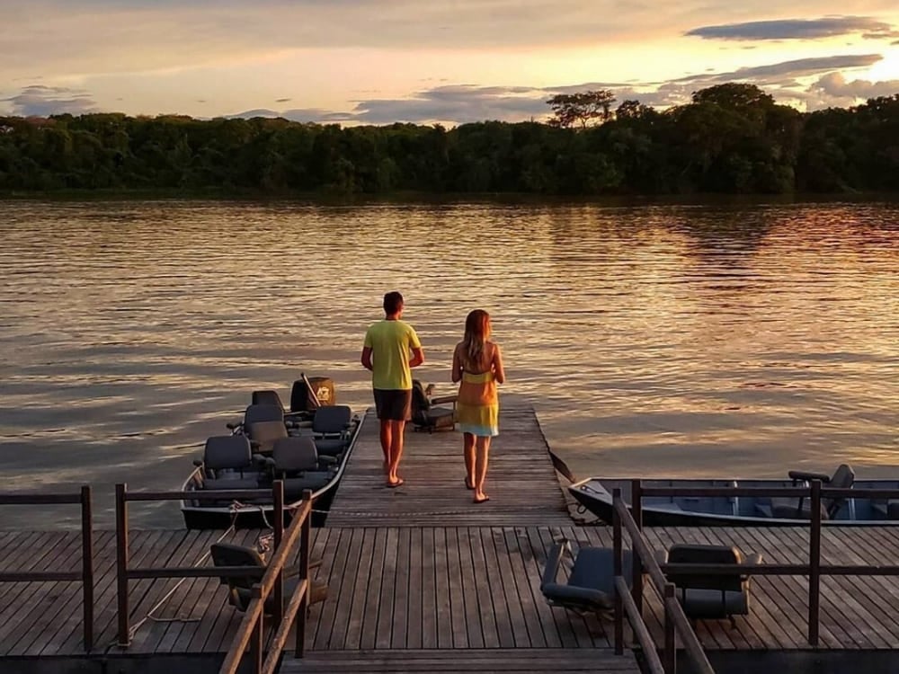 A couple walks down a wooden pier toward safari boats during a beautiful sunset over a Pantanal river.