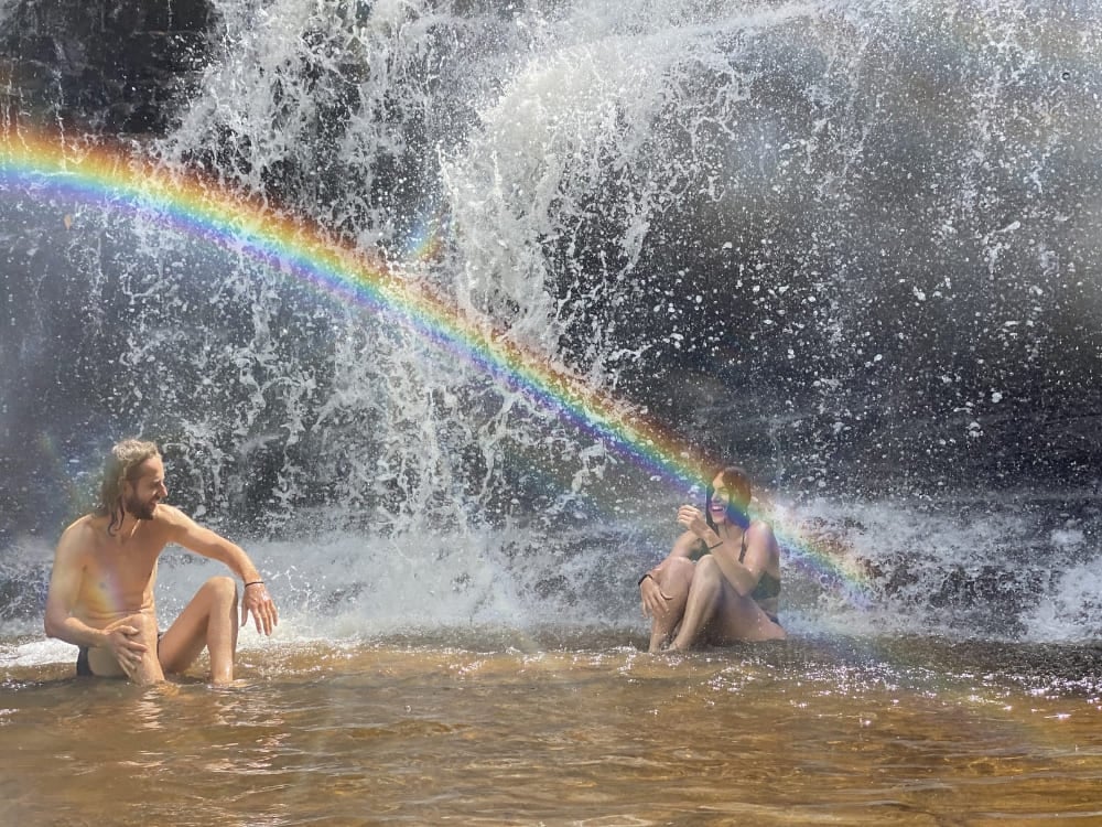 Two travelers sitting in the shallow water at the base of a waterfall with a rainbow reflection.