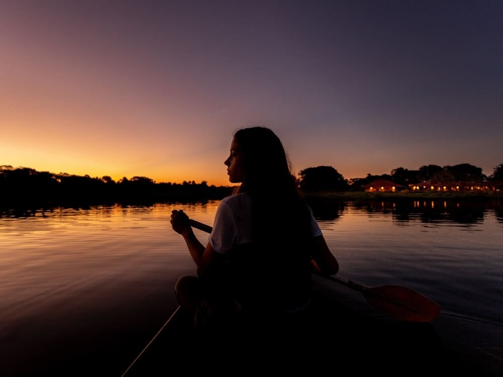 Silhouette of a person paddling a canoe on the calm waters of a river during a peaceful sunset.