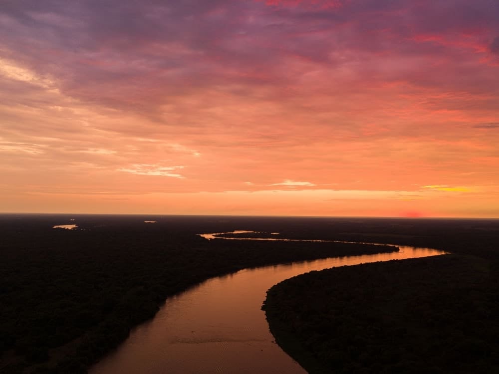 Aerial view of a winding river flowing through the vast wetlands, reflecting the pink and orange evening sky.