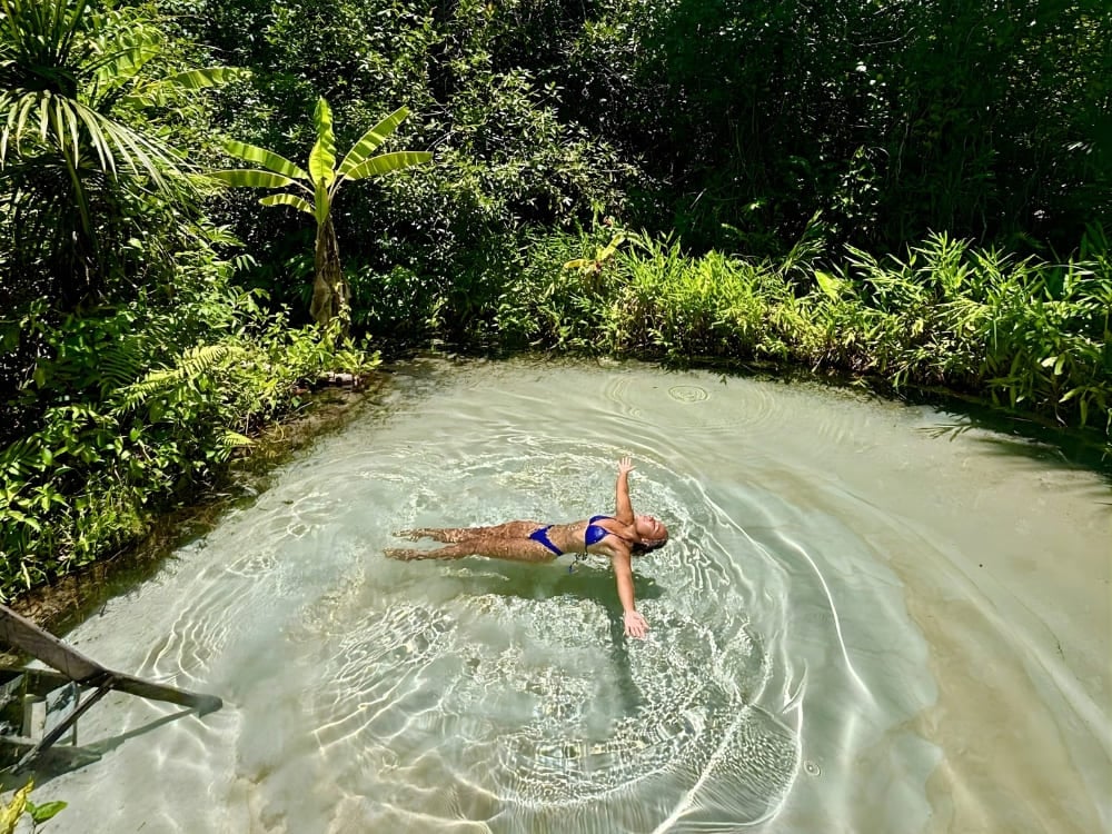 Woman floating on her back with her arms spread in crystal clear light blue waters, surrounded by tropical trees and dense green vegetation.