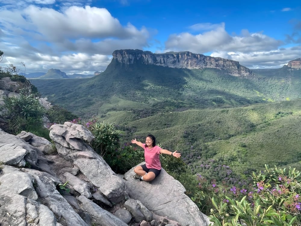 Smiling woman sitting cross-legged with open arms on a rock, admiring a green valley with an imposing flat-topped mountain in the background.
