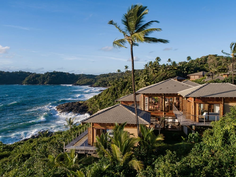 Luxurious villas with wooden roofs surrounded by palm trees on a cliff facing the sea.