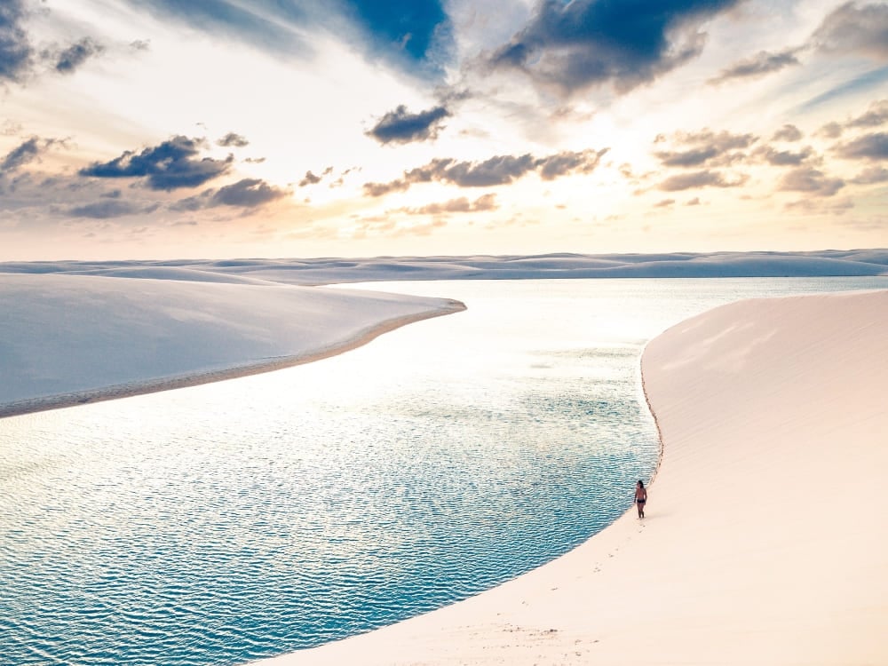 A single person walks along the edge of a vibrant blue lagoon winding through vast white sand dunes under a dramatic cloudy sky.