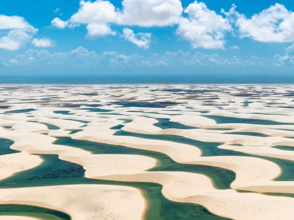 An aerial view of a vast expanse of white sand dunes interspersed with vibrant blue and green seasonal lagoons under a bright blue sky with fluffy white clouds in Lençóis Maranhenses National Park, Brazil.