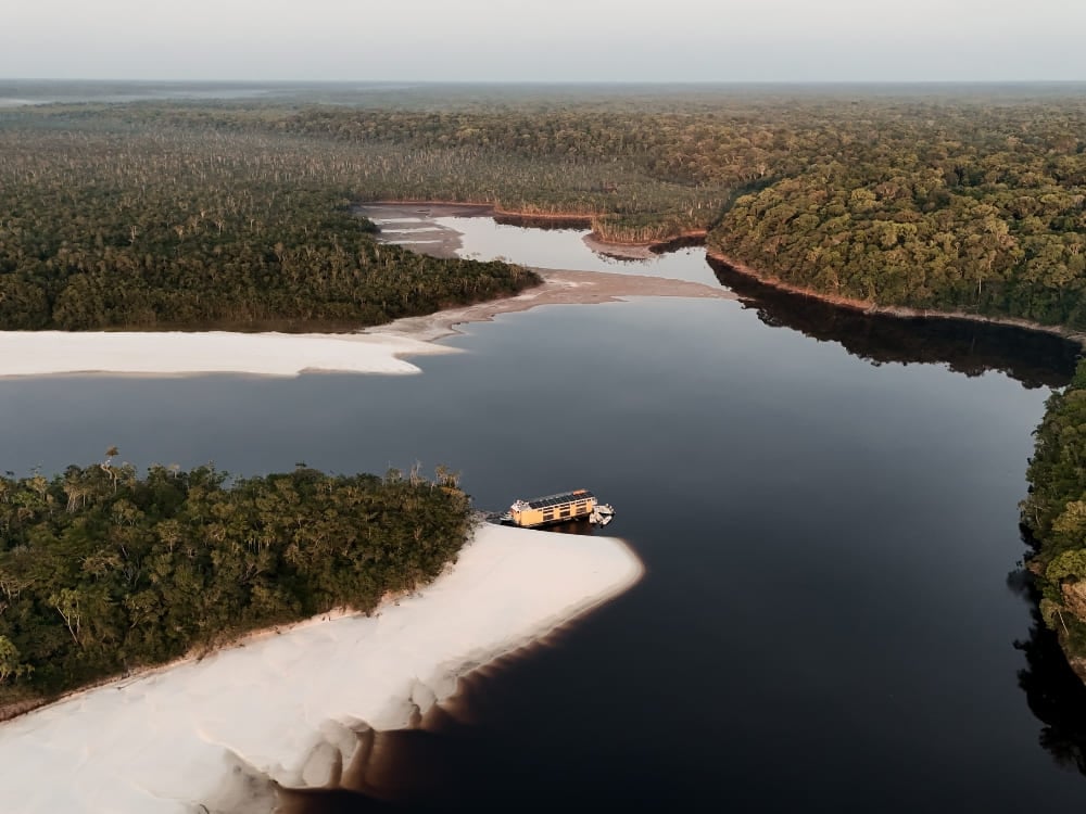 An aerial view of a small riverboat moored on a white sandbank beside a dark Amazon river, surrounded by rainforest.