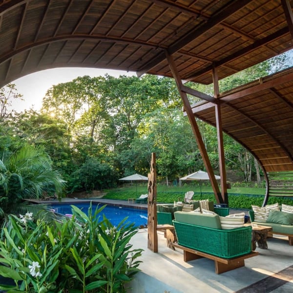 A covered lounge deck with green sofas beside a swimming pool at sunset, framed by tropical plants and trees at an Amazon ecolodge.