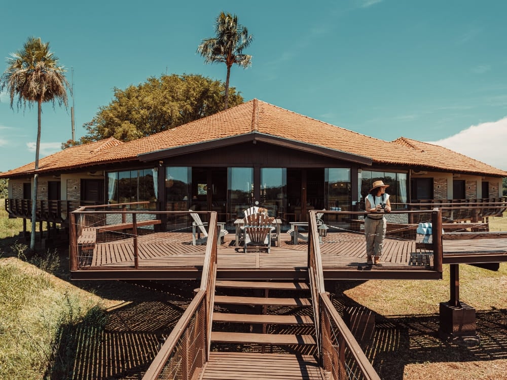 The main building of a Pantanal wildlife lodge with a tiled roof and a wide wooden deck under a bright blue sky, a guest standing between palm trees.