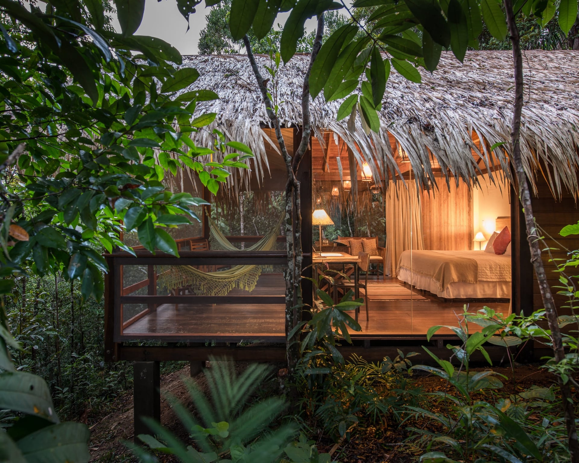 A luxury Amazon jungle lodge bungalow with a thatched roof and glass walls glowing with warm light at dusk, a hammock on its wooden deck amid the rainforest.