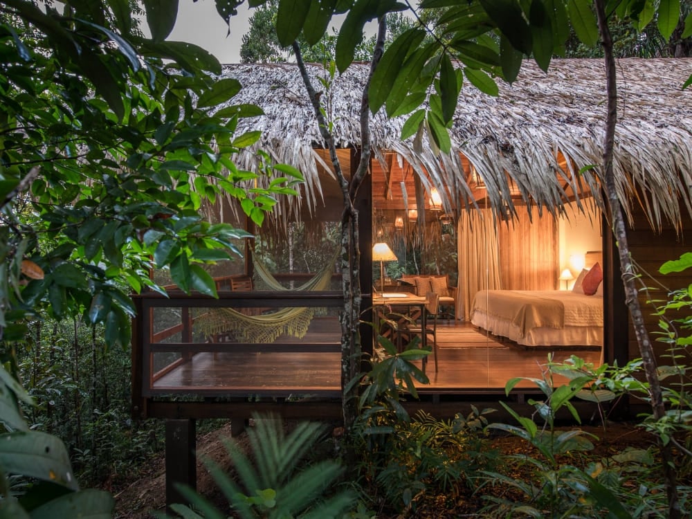 A luxury Amazon lodge bungalow with a thatched roof and glass walls glowing with warm light at dusk, a hammock on its deck amid the rainforest.