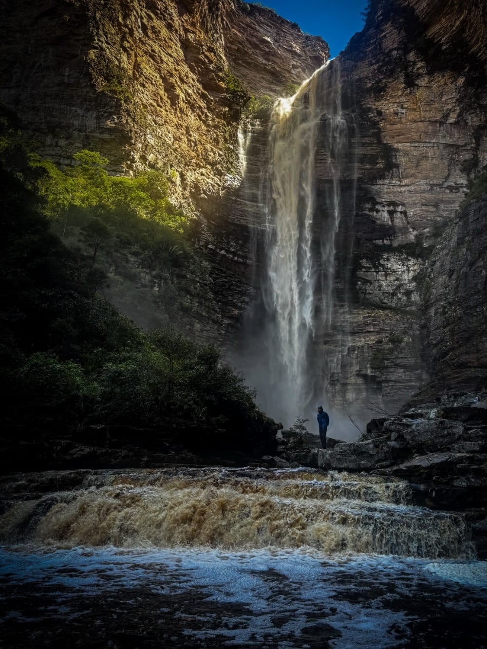 A lone hiker at the base of the Cachoeira da Encantada, falling between the canyon walls above the amber Rio Samina, Chapada Diamantina, Brazil.