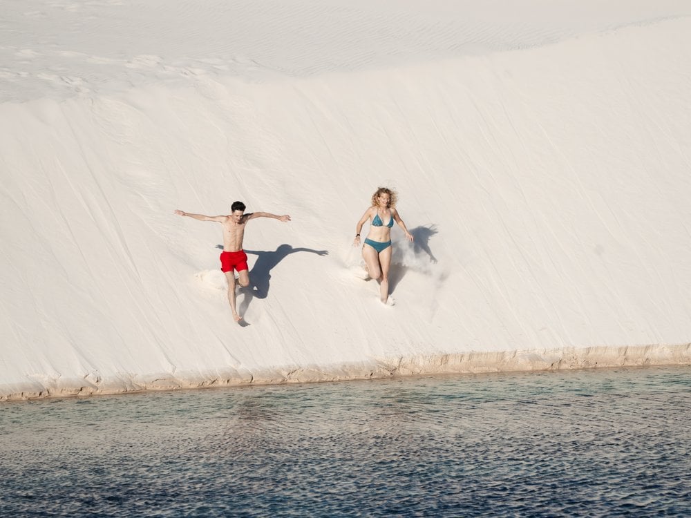 Two travelers running down a white dune toward a lagoon in Lençóis Maranhenses