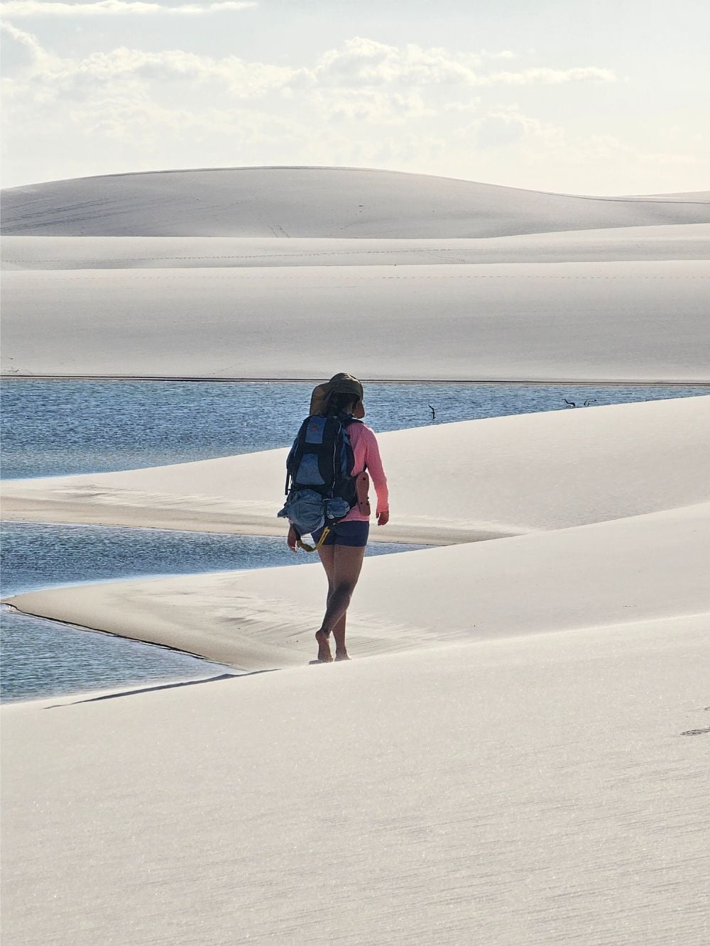 A backpacker walks barefoot along the white sand dunes near a blue rainwater lagoon while trekking in Lençóis Maranhenses National Park. A backpacker walks barefoot along the white sand dunes near a blue rainwater lagoon while trekking in Lençóis Maranhenses National Park.