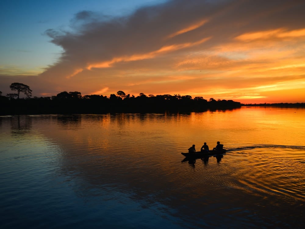 Silhouette of three people in a small canoe navigating the Amazon River against a bright orange sunset sky. Amazon Rainforest travel Silhouette of three people in a small canoe navigating the Amazon River against a bright orange sunset sky.