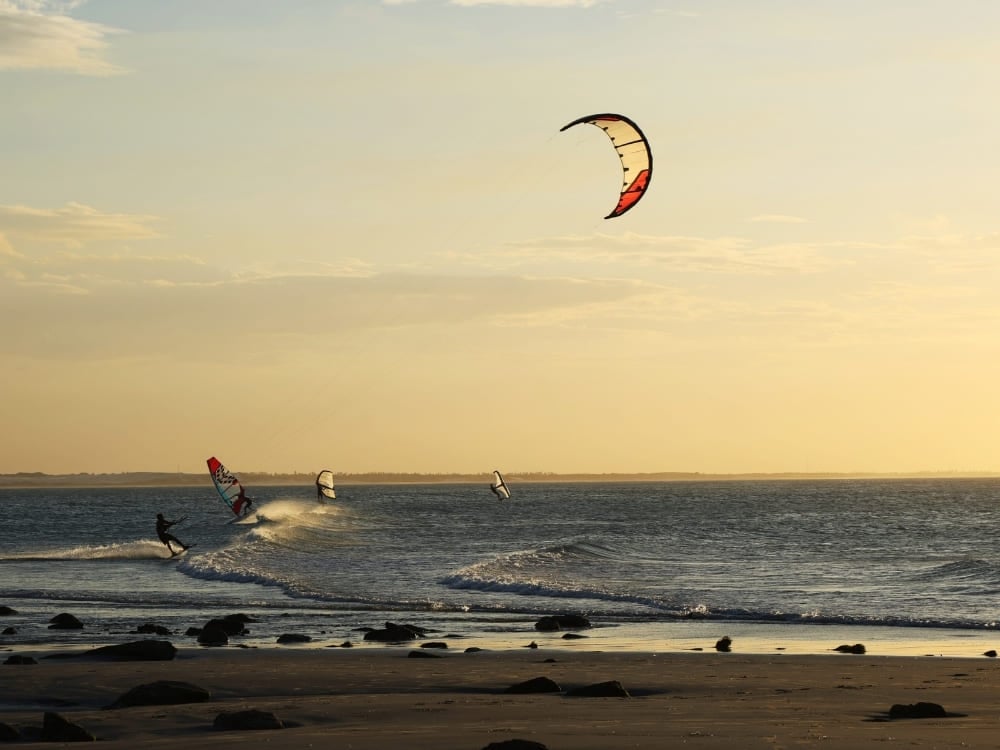 . Silhouettes of people kitesurfing and windsurfing on ocean waves near a sandy beach during a golden sunset.