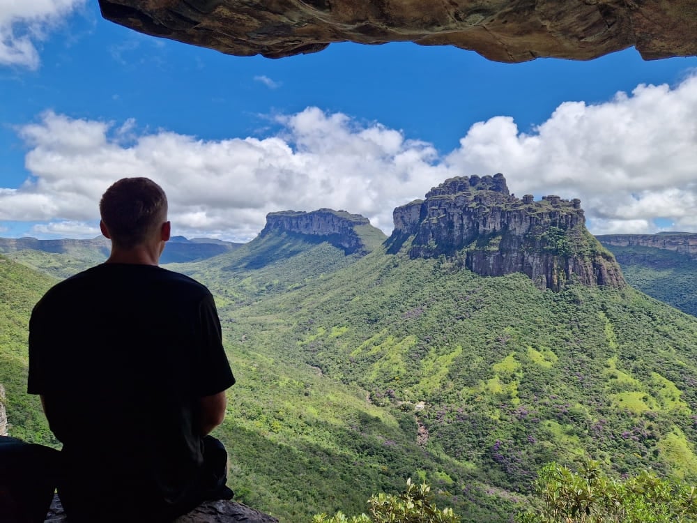 a man sitting under a rocky overhang observing a vast green valley and flat-topped mountains in Chapada Diamantina. a man sitting under a rocky overhang observing a vast green valley and flat-topped mountains in Chapada Diamantina.