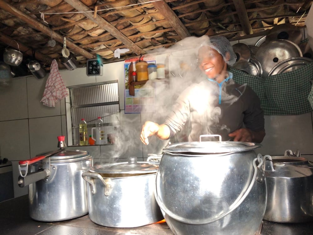 A local woman smiling as she cooks a traditional homemade meal using large aluminum pots over a stove. A local woman smiling as she cooks a traditional homemade meal using large aluminum pots over a stove.
