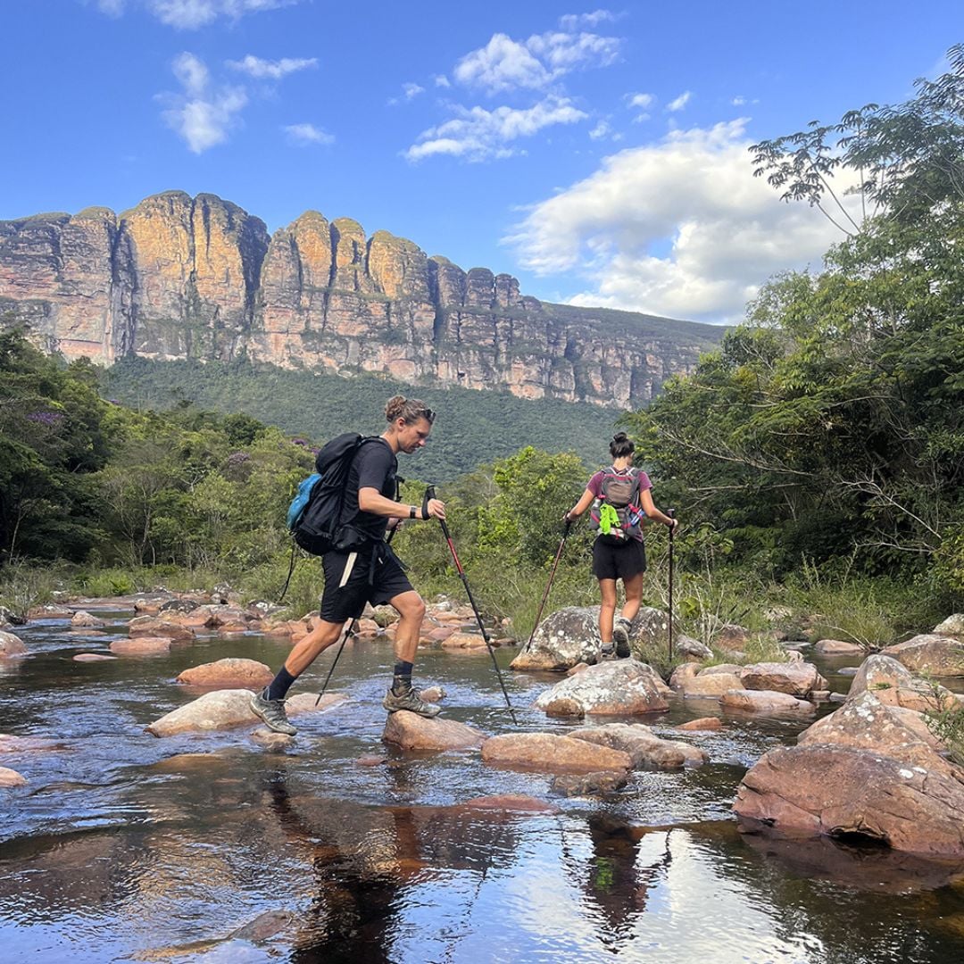 Two hikers using trekking poles to cross a shallow, rocky river with a large mountain peak towering in the background. Two hikers using trekking poles to cross a shallow, rocky river with a large mountain peak towering in the background.