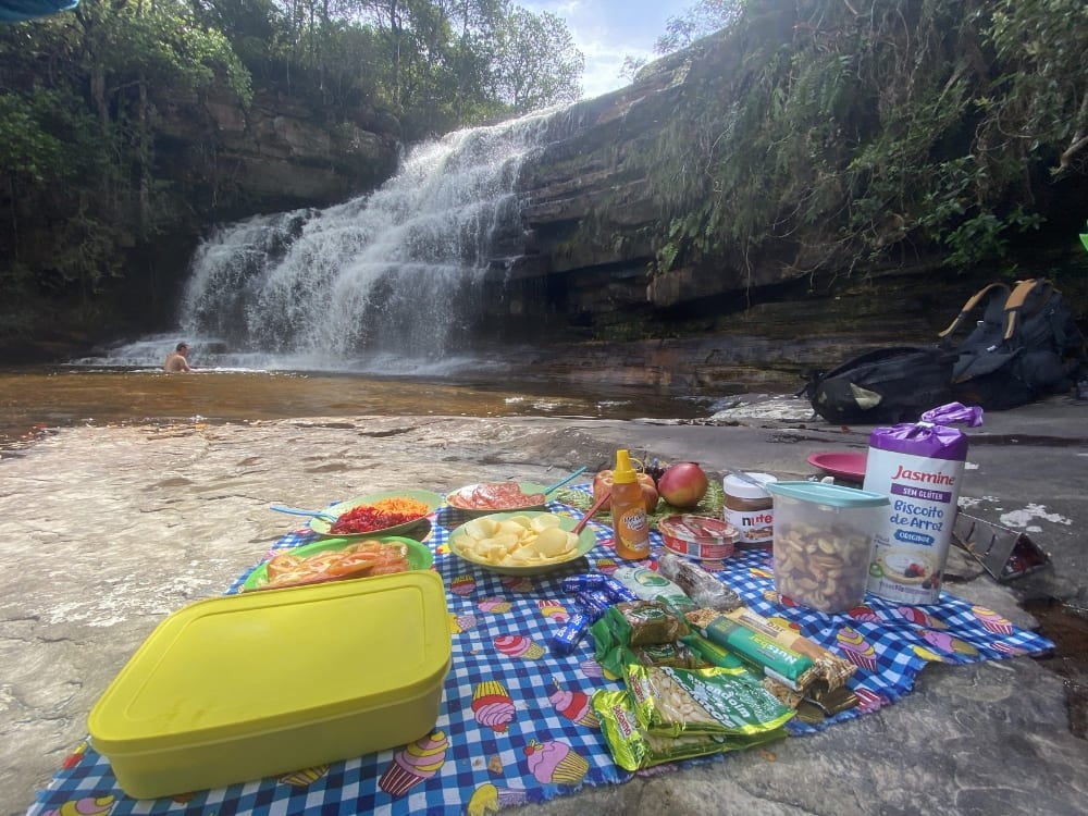 A colorful picnic spread with fruits, snacks, and sandwiches laid out on a blanket beside a small cascading waterfall. A colorful picnic spread with fruits, snacks, and sandwiches laid out on a blanket beside a small cascading waterfall.