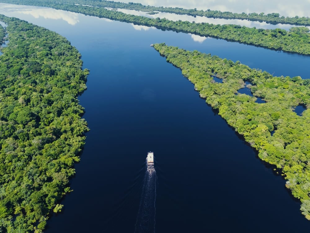 A multi-level riverboat travels along a wide, calm dark river bordered by dense green rainforest islands reflecting the sky. A multi-level riverboat travels along a wide, calm dark river bordered by dense green rainforest islands reflecting the sky.