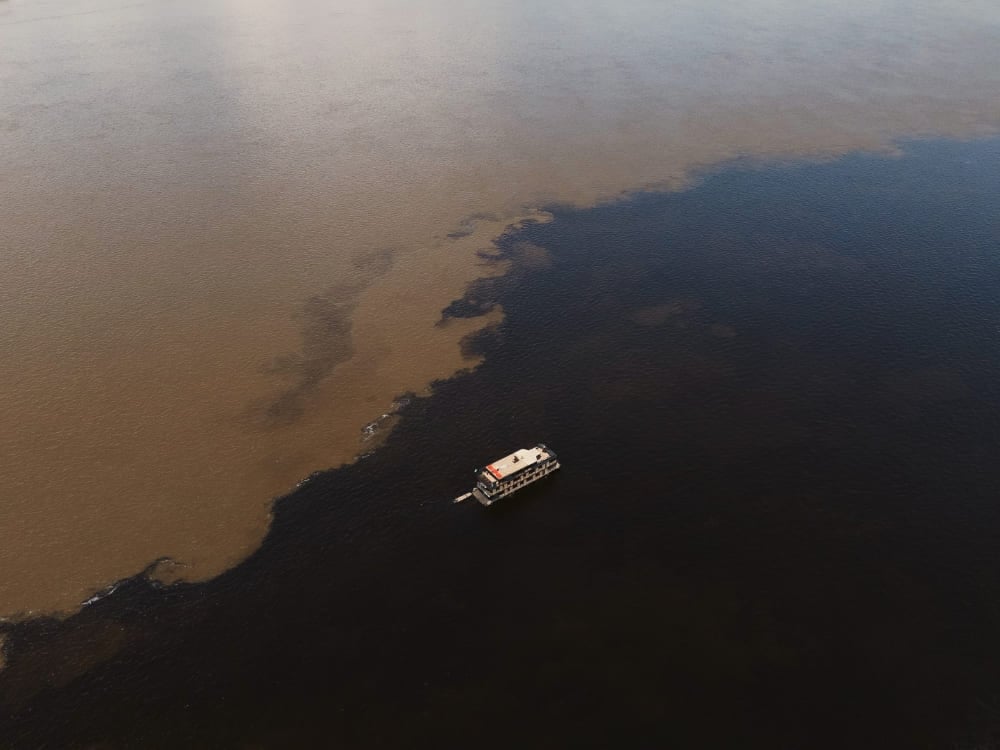 Aerial view of a riverboat navigating the Meeting of the Waters in the Amazon, showing the distinct dark and sandy rivers flowing side-by-side. Aerial view of a riverboat navigating the Meeting of the Waters in the Amazon, showing the distinct dark and sandy rivers flowing side-by-side.
