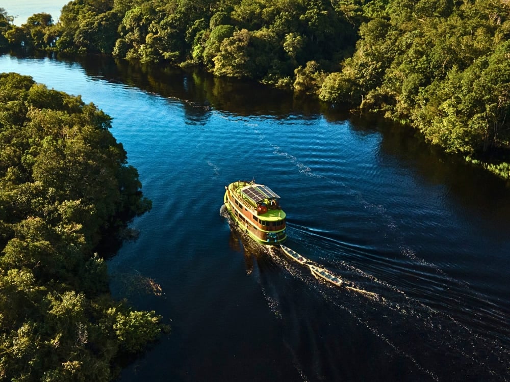 A green ecotourism riverboat with solar panels cruises down a dark river surrounded by lush Amazon rainforest. A green ecotourism riverboat with solar panels cruises down a dark river surrounded by lush Amazon rainforest.