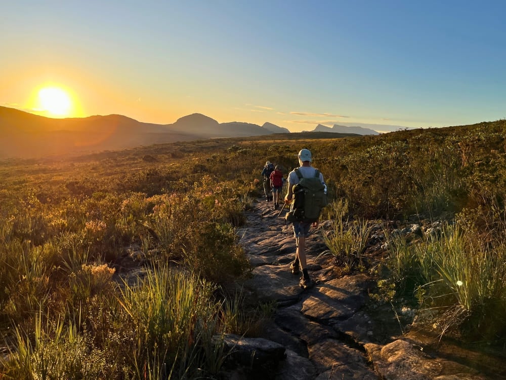 Hikers walk along a rocky trail towards distant mountains during a golden sunset in Vale do Pati, Chapada Diamantina National Park. Hikers walk along a rocky trail towards distant mountains during a golden sunset in Vale do Pati, Chapada Diamantina National Park.
