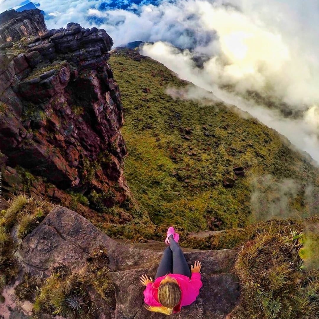 A hiker sits on the edge of a steep, rocky sandstone cliff on Mount Roraima, looking down at a vast valley filled with thick white clouds. A hiker sits on the edge of a steep, rocky sandstone cliff on Mount Roraima, looking down at a vast valley filled with thick white clouds.