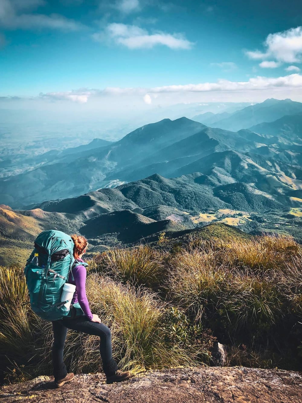 A hiker wearing a large green backpack stands on a grassy mountain ridge, overlooking the expansive, mountainous landscape of Serra Fina. A hiker wearing a large green backpack stands on a grassy mountain ridge, overlooking the expansive, mountainous landscape of Serra Fina.