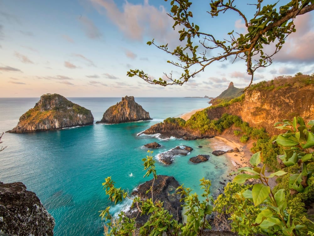 The iconic Morro dos Dois Irmãos rock formations rise from the turquoise ocean along a lush, cliff-lined beach in Fernando de Noronha. The iconic Morro dos Dois Irmãos rock formations rise from the turquoise ocean along a lush, cliff-lined beach in Fernando de Noronha.