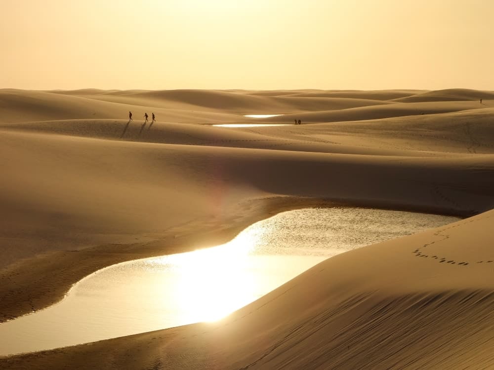 Travelers walk across the vast expanse of sand dunes in Lençóis Maranhenses during a bright sunset. Travelers walk across the vast expanse of sand dunes in Lençóis Maranhenses during a bright sunset.