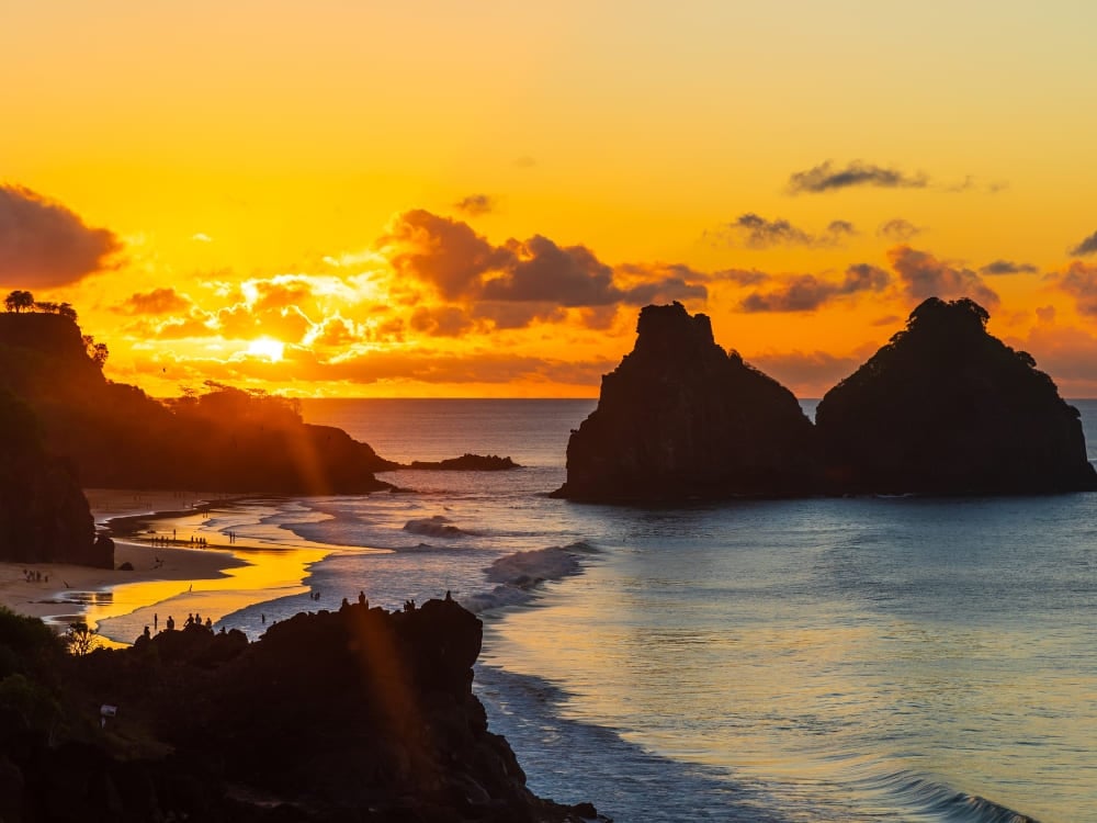 A golden sunset illuminates the ocean waves and the Morro dos Dois Irmãos rock formations as surfers paddle in the water. A golden sunset illuminates the ocean waves and the Morro dos Dois Irmãos rock formations as surfers paddle in the water.