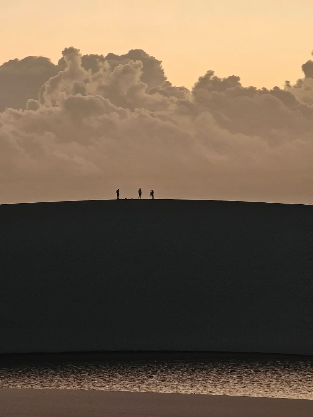 Silhouettes of three people walking on top of a sand dune in Lencois Maranhenses during sunrise, with clouds in the background. Silhouettes of three people walking on top of a sand dune in Lencois Maranhenses during sunrise, with clouds in the background.