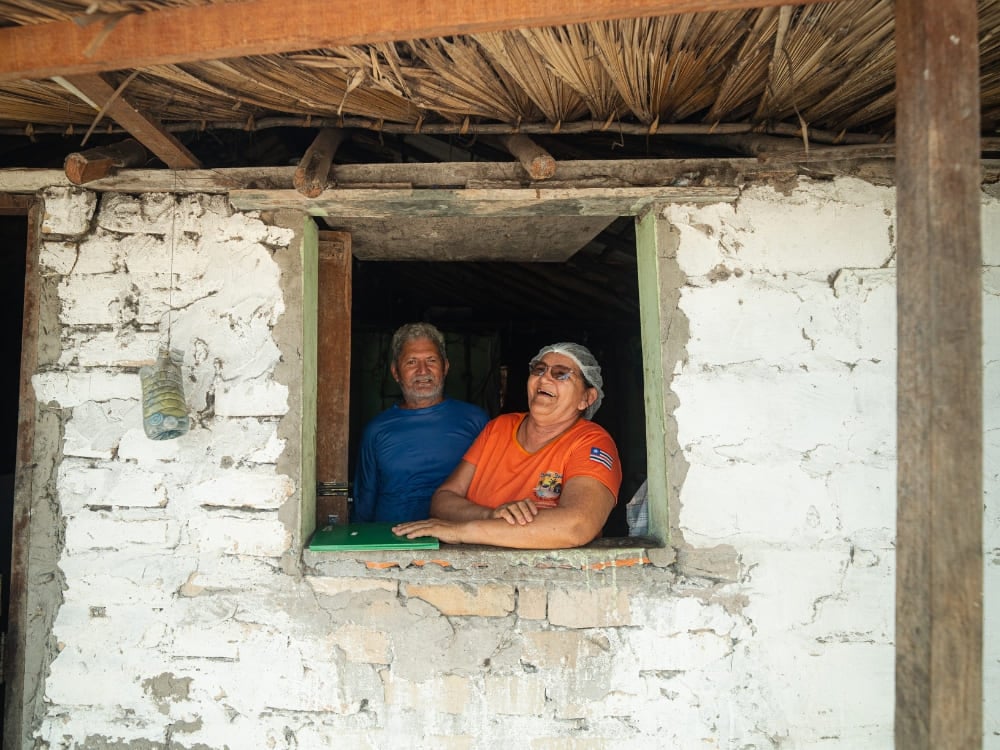 A smiling older man and woman look out the window of a rustic, white-washed house with a thatched palm roof in Lençóis Maranhenses. A smiling older man and woman look out the window of a rustic, white-washed house with a thatched palm roof in Lençóis Maranhenses.