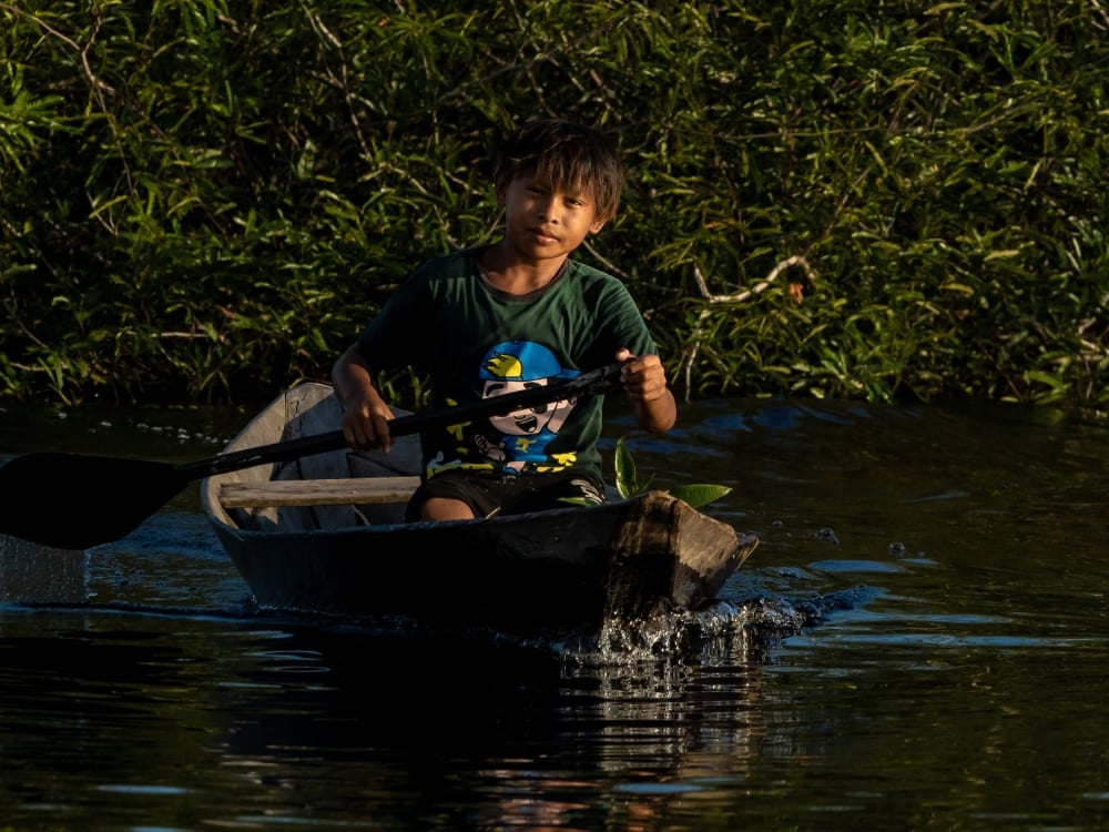A young boy paddling a wooden canoe on a dark river, representing the daily life of riverside communities in the Amazon. A young boy paddling a wooden canoe on a dark river, representing the daily life of riverside communities in the Amazon.