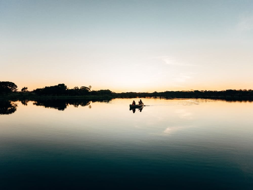 Two people paddling a canoe on a calm river that mirrors the sky in the area where is the Pantanal. Two people paddling a canoe on a calm river that mirrors the sky in the area where is the Pantanal.